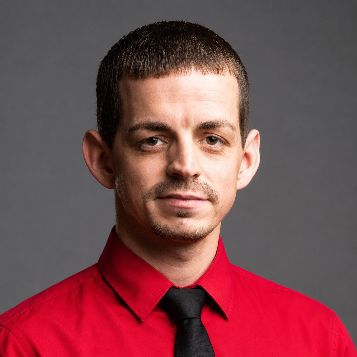 Professional headshot of Josh Centanni, an author and speaker, wearing a red dress shirt with a black necktie against a solid dark gray studio background.