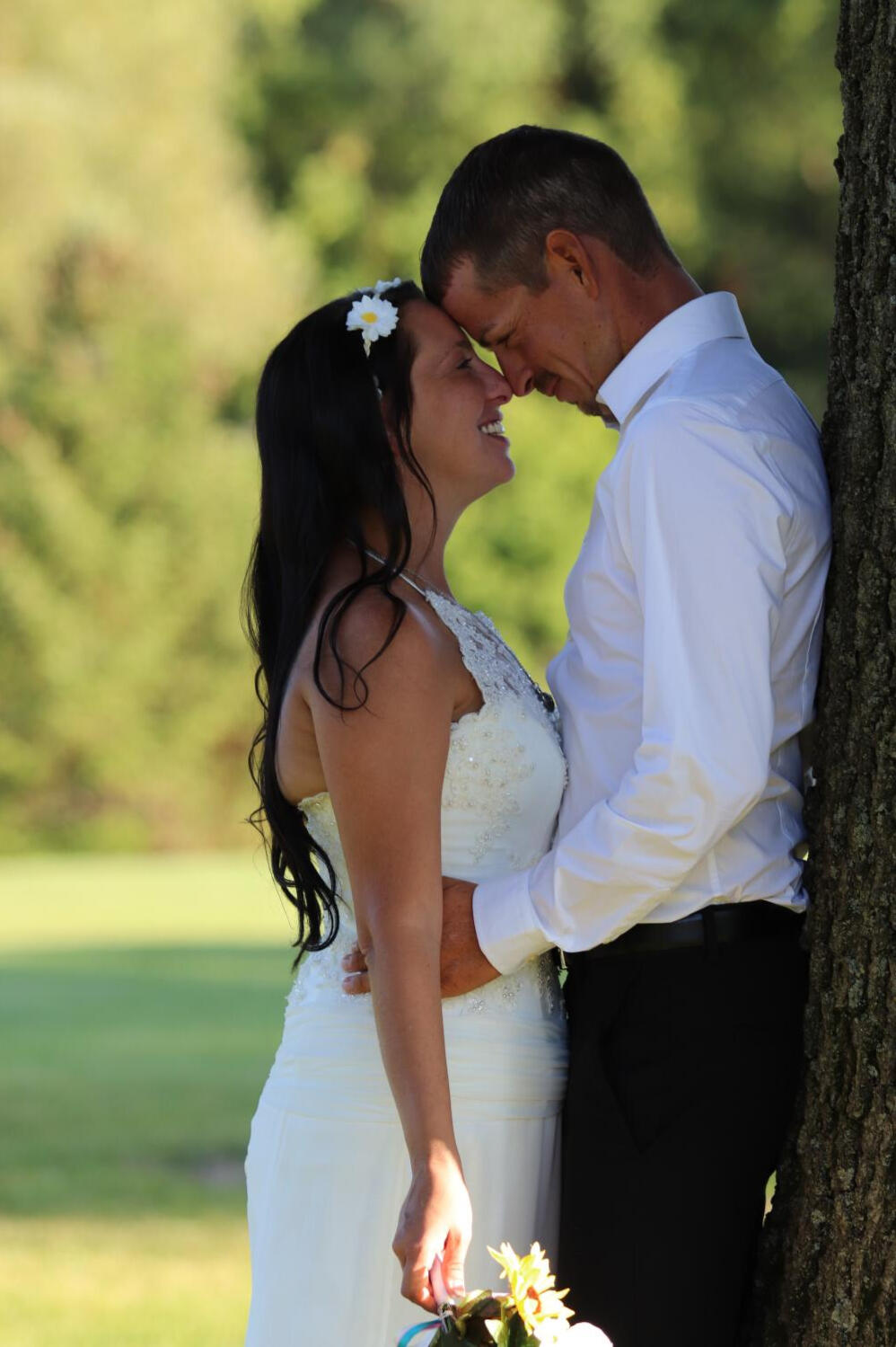 Close-up portrait of a bride and groom smiling warmly at each other with their foreheads together under a tree, with a soft blurred green grass background.